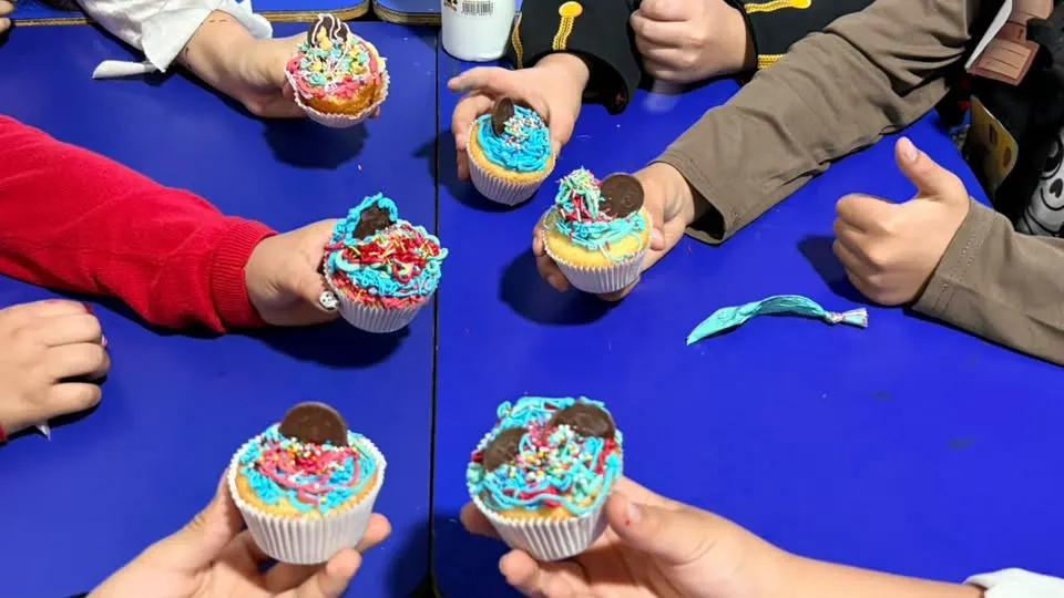 Manos de estudiantes sostienen cupcakes decorados sobre una mesa azul
